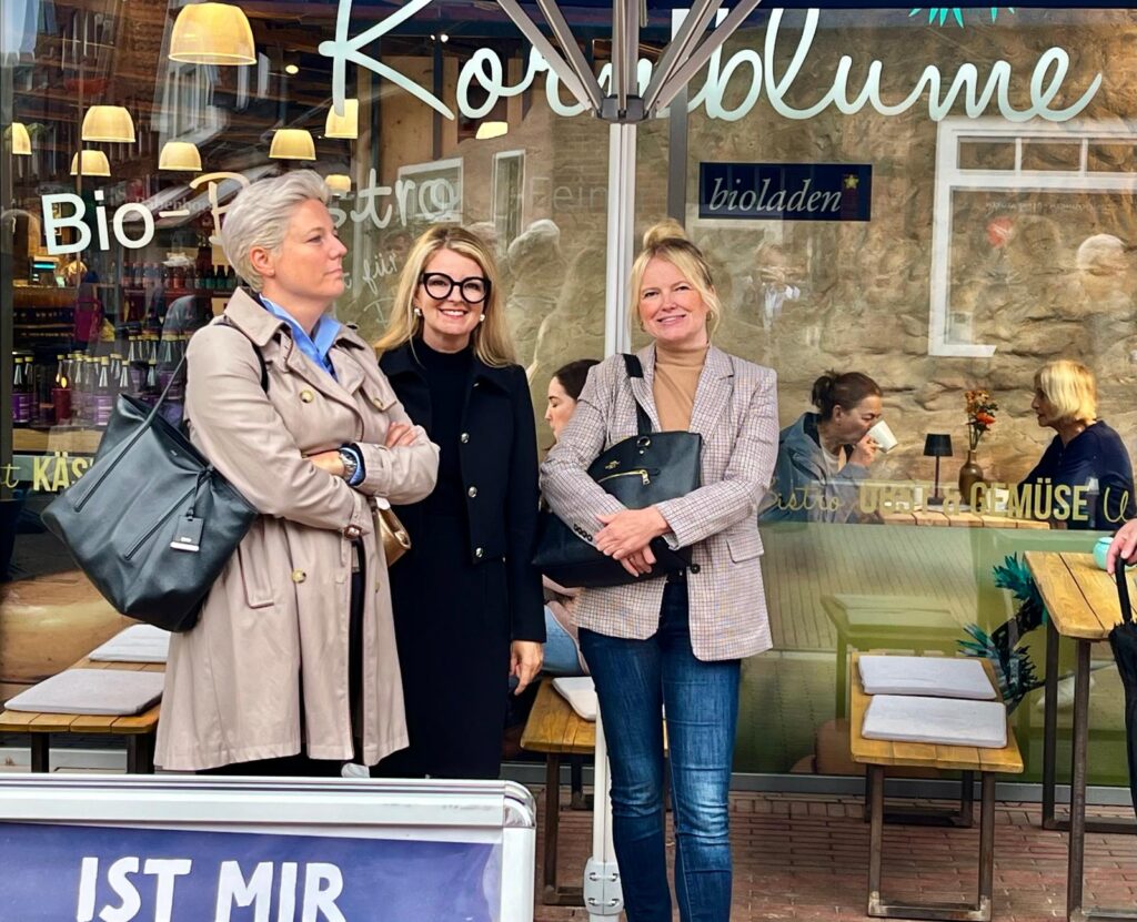 Drei Frauen stehen beim Jurybesuch vor dem Schaufenster des Bioladens Kornblume.